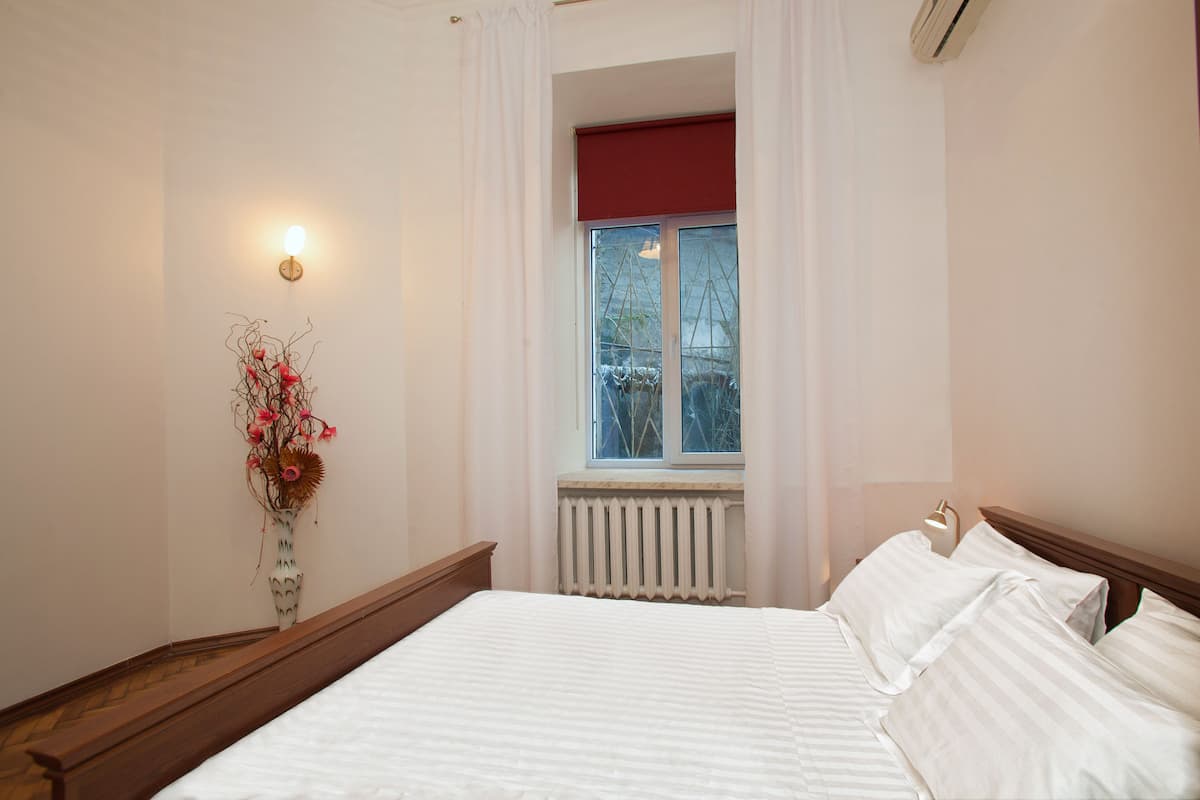 A bedroom with a bed, a radiator, a window with curtains, and a wall-mounted light fixture.