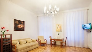 Una sala de estar con sofá, sillas, una mesa, una araña, un cuadro, televisor y una ventana con cortinas.