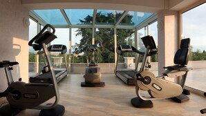 A gym with three stationary bikes, a treadmill, and a rowing machine, with large windows offering a view of trees and buildings. - Palazzo Giordano Bruno (Nola)