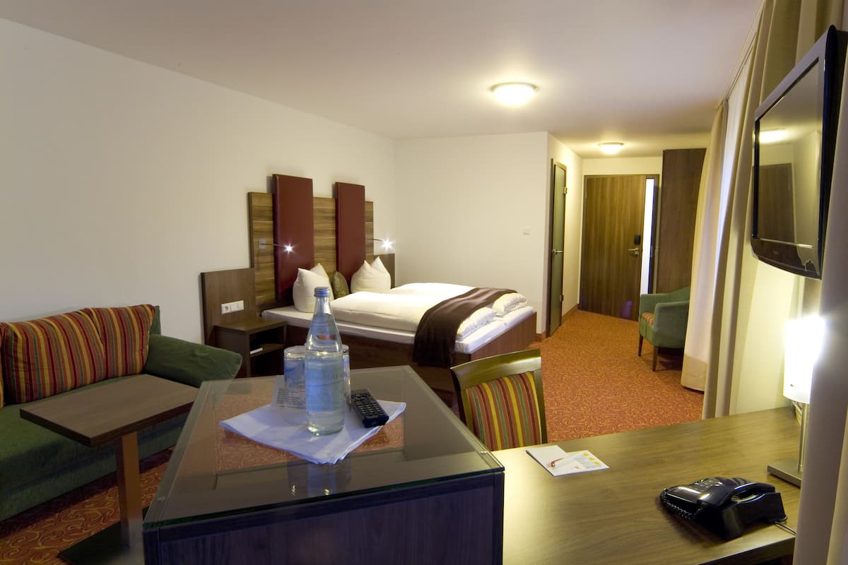A hotel room with a bed, a sofa, a desk with a glass top, a chair, a TV, and a bedside table.