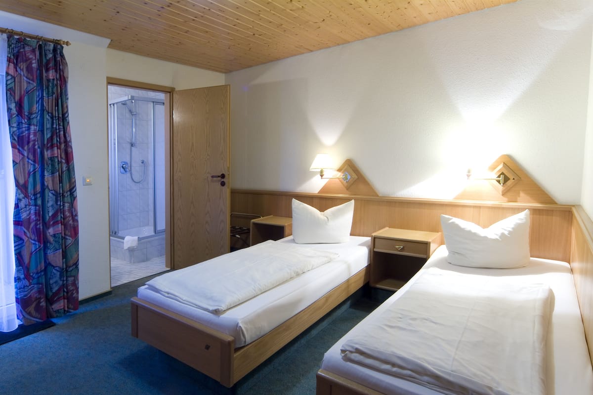 A hotel room with two single beds, a shower, wooden headboard, and a small bedside table with a lamp.