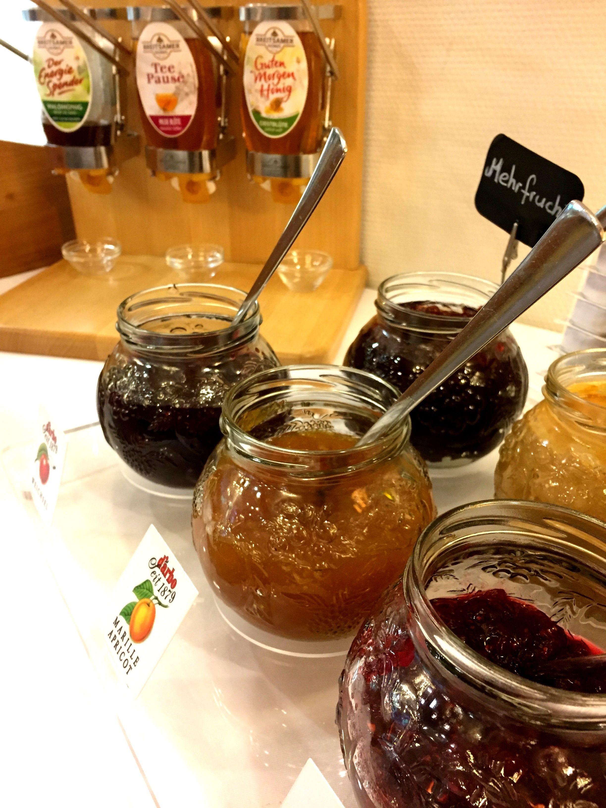 En uppvisning av olika sorters marmelad och honung i glasburkar, med metallskedar, och vissa burkar var märkta med namn som "Marillen Apricot" och "Tee Pause".
