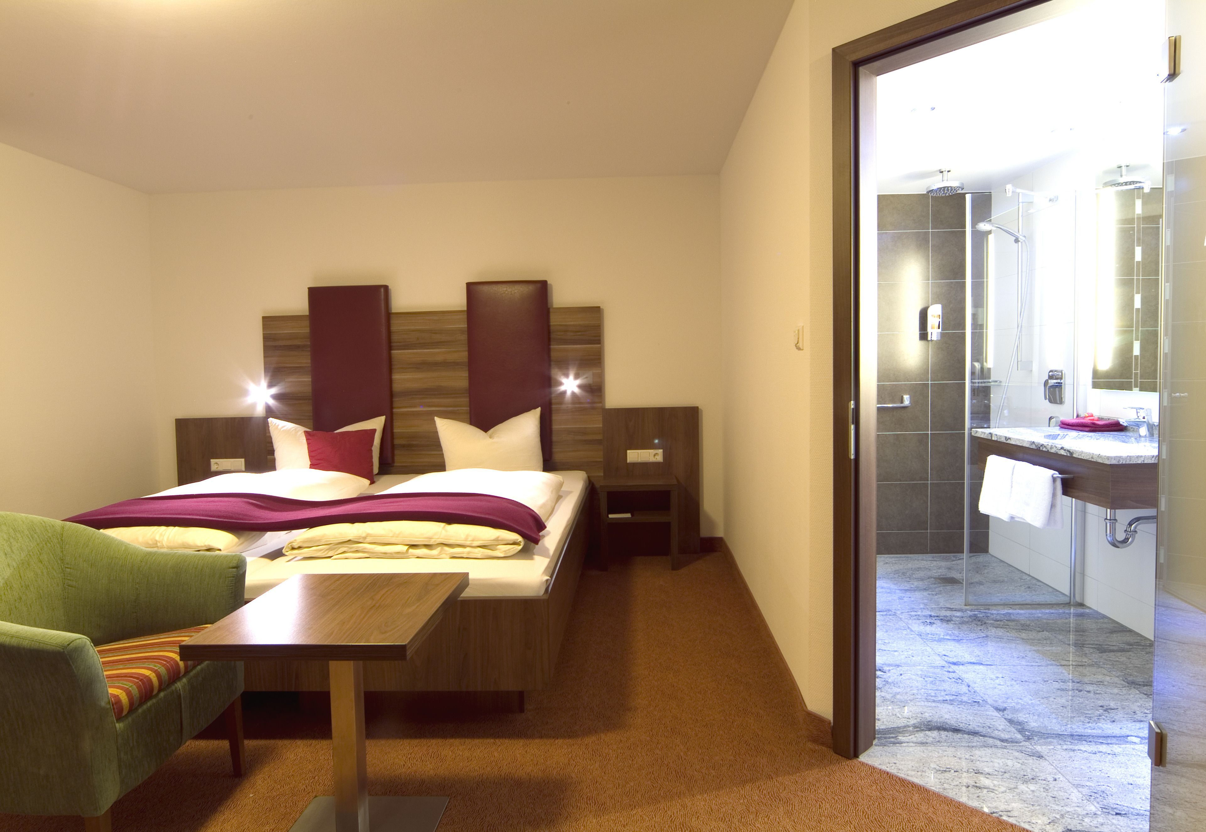 A hotel room with a bed, bedside table, chair, and a bathroom visible through an open door.