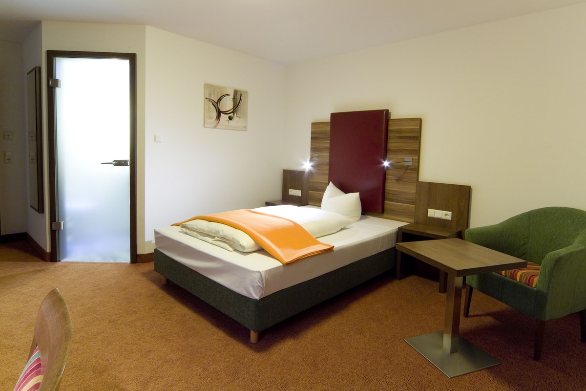 A hotel room with a bed, a green armchair, a small table, and a painting on the wall.