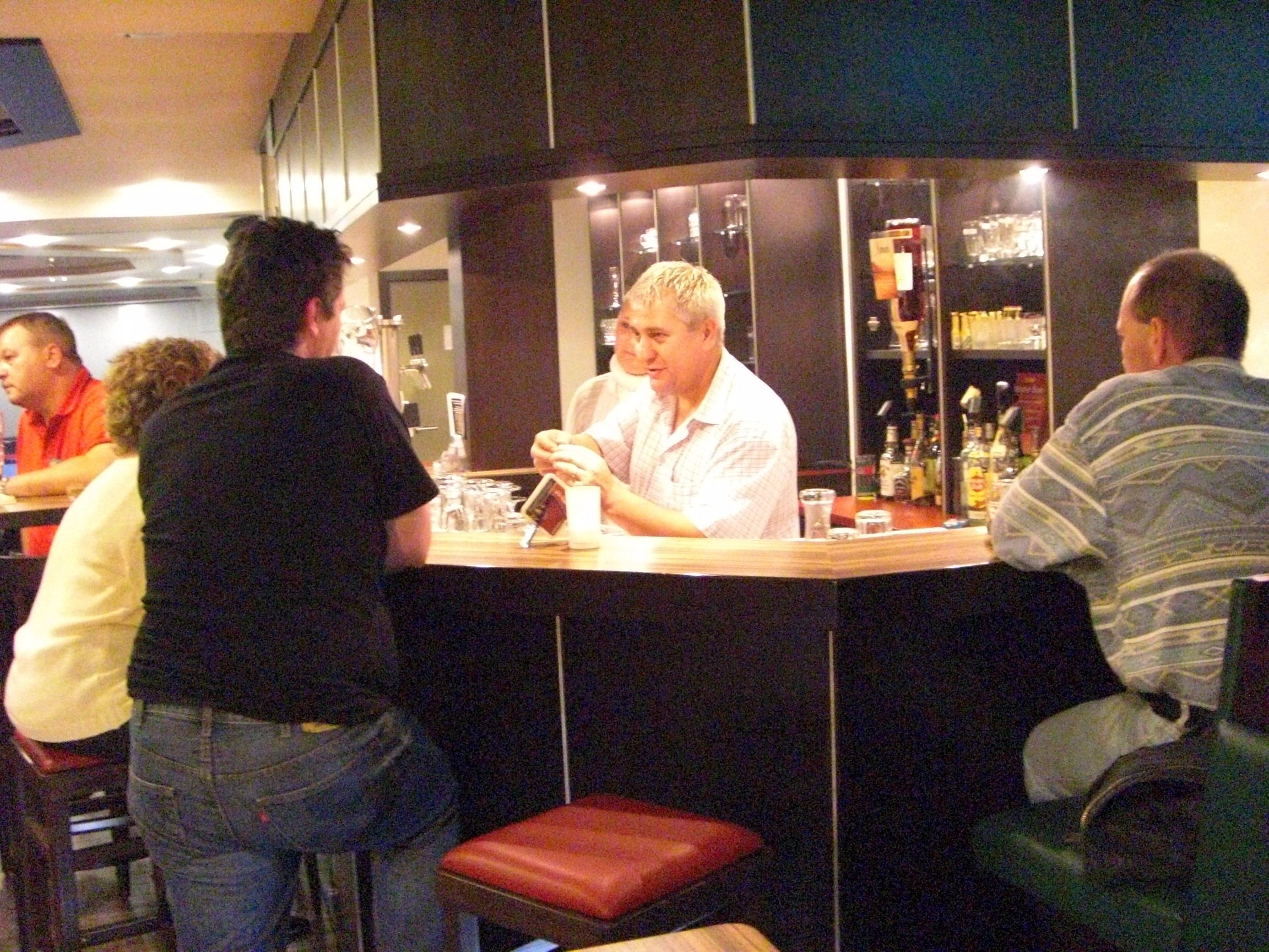 Een bar met een toonbank, krukken en een plank met flessen.