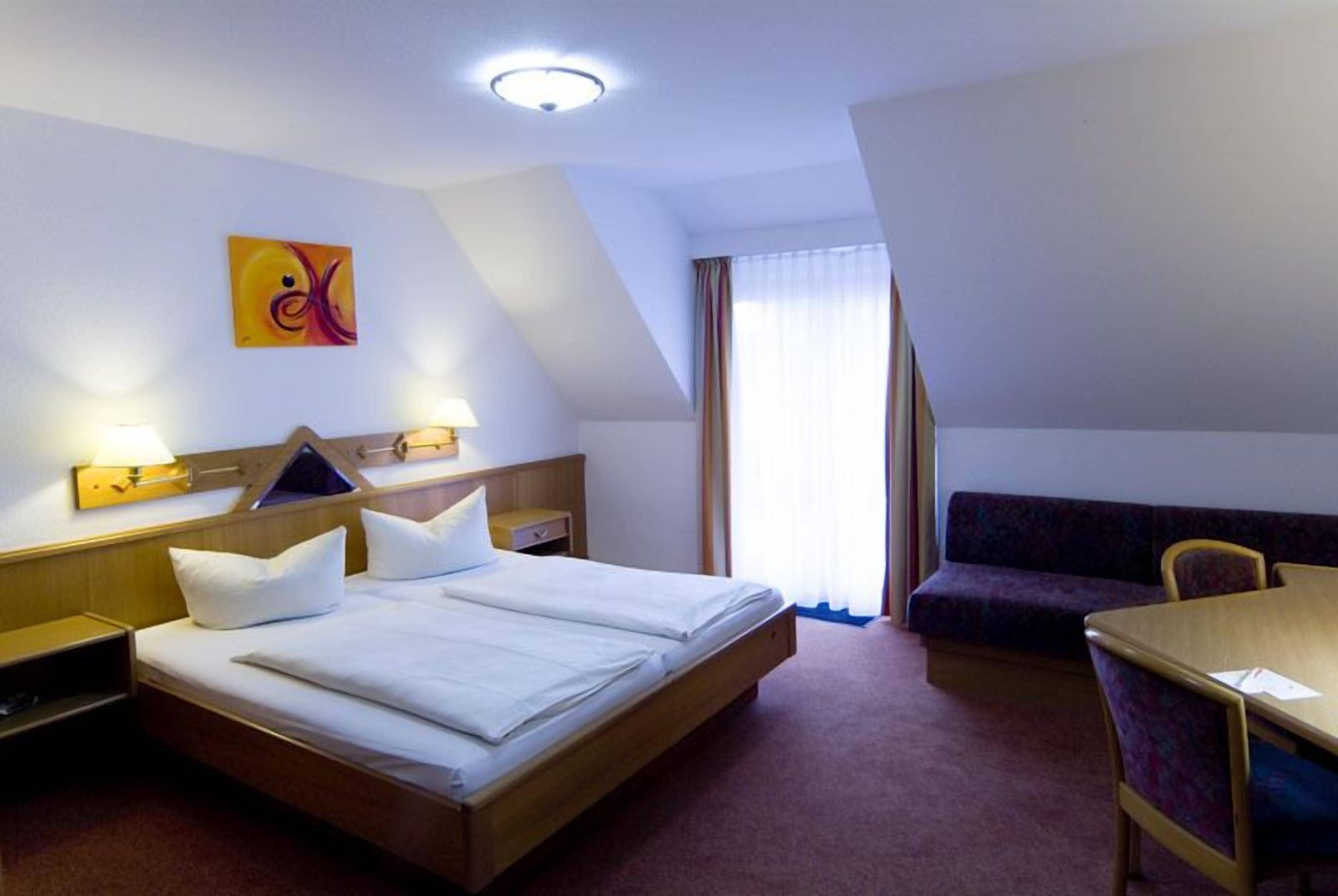 A hotel room with a bed, bedside table, lamp, artwork, a sofa, and a table.