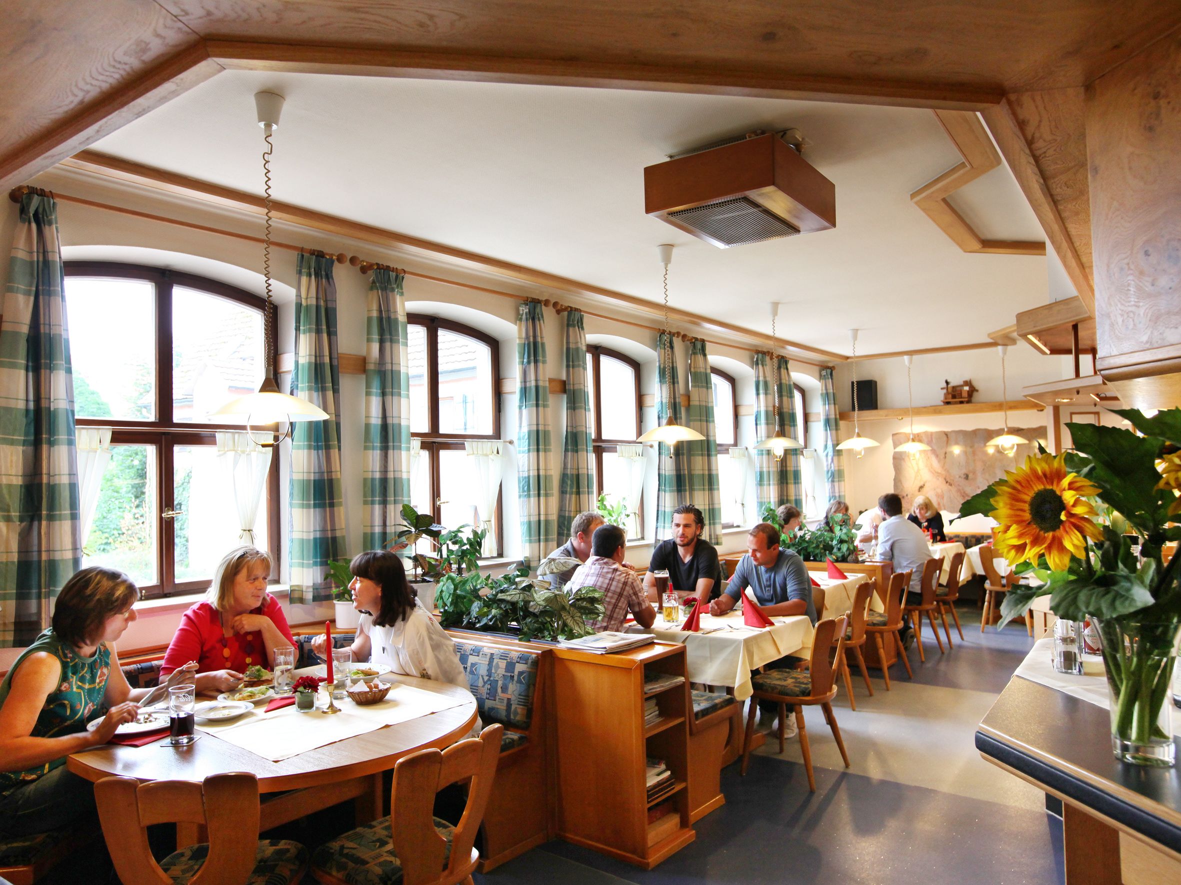 Un restaurante con mesas redondas, sillas de madera y ventanas amplias con cortinas.