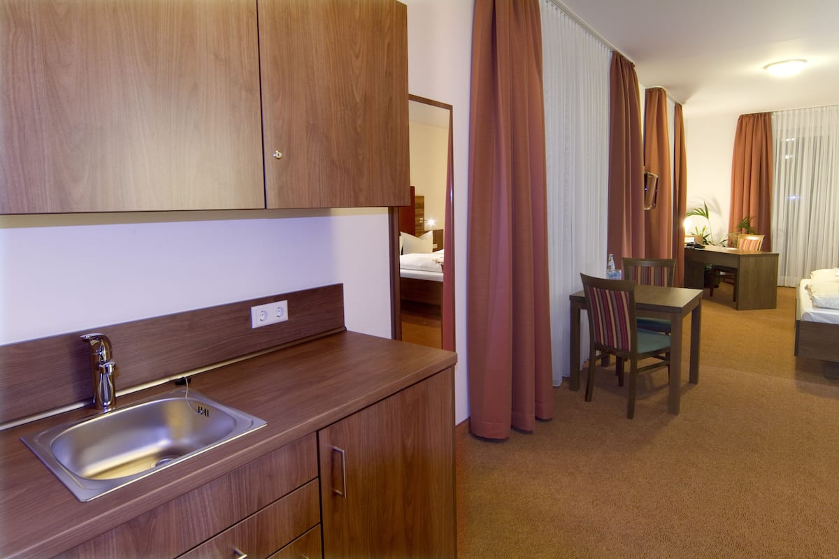 A hotel room with a wooden kitchenette, a bed, a desk, and a chair.