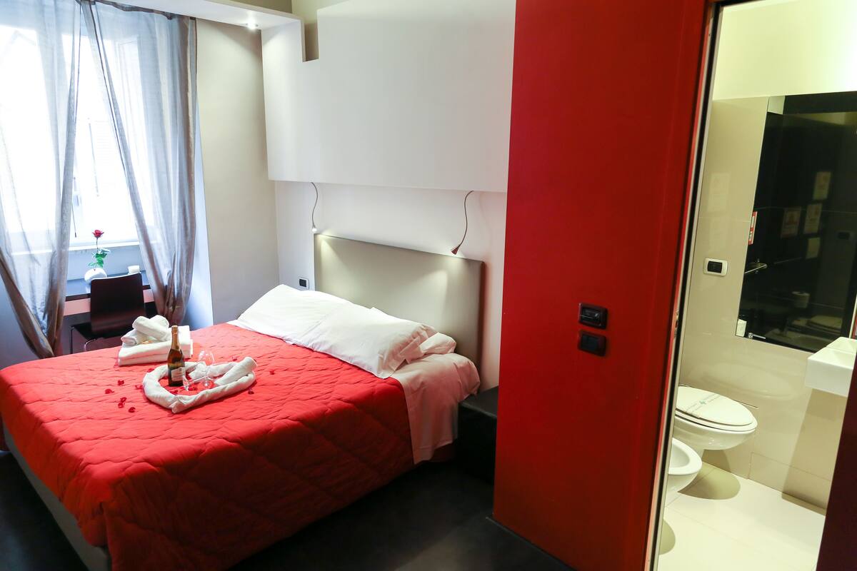 Una habitación de hotel con una colcha roja, una cama bien hecha con sábanas blancas, una botella de champán y una copa sobre la cama.