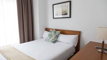 A neatly made bed with white linens and a single decorative pillow, a wooden headboard, a framed picture on the wall, and a bedside table with a lamp.