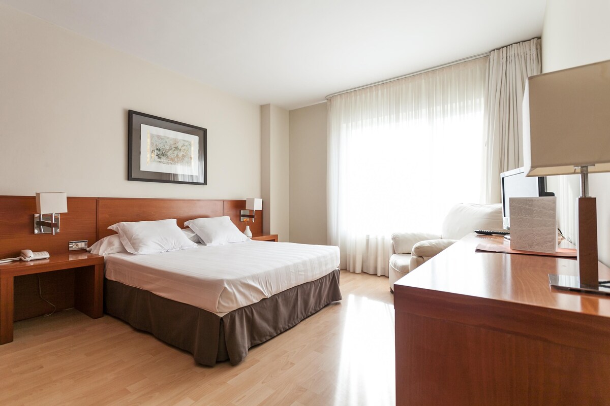 A hotel room with a large bed, a desk, a chair, a lamp, and a window with curtains.