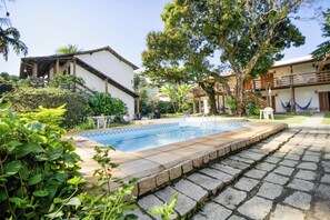 Una piscina rodeada de baldosas de piedra, con un edificio blanco y una hamaca al fondo.