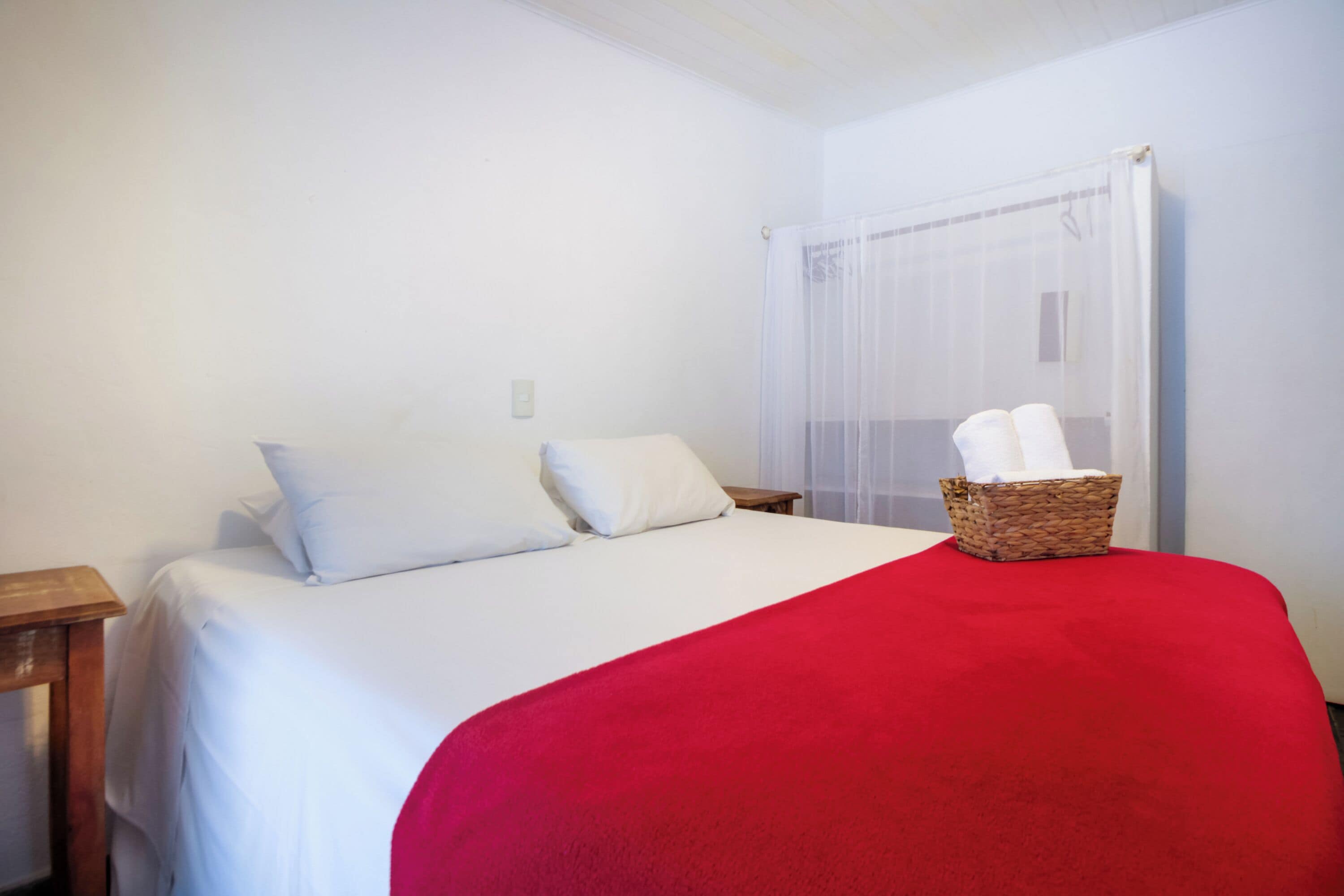 A neatly made bed with white linens and a red blanket, a wooden nightstand, and a wicker basket with towels on the bed.