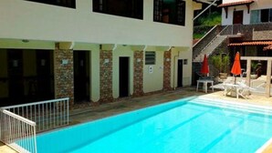 Una piscina con agua cristalina azul, rodeada por un edificio blanco con varias puertas y ventanas. Hay dos sombrillas rojas y sillas reclinables blancas cerca de la piscina.