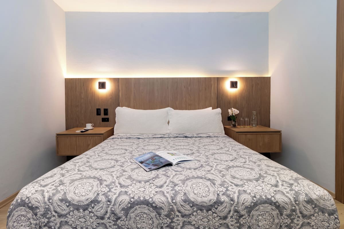 Un lit bien fait, recouvert d’une couvre-lit à motifs, deux tables de chevet avec des lampes et un luminaire fixé au mur.