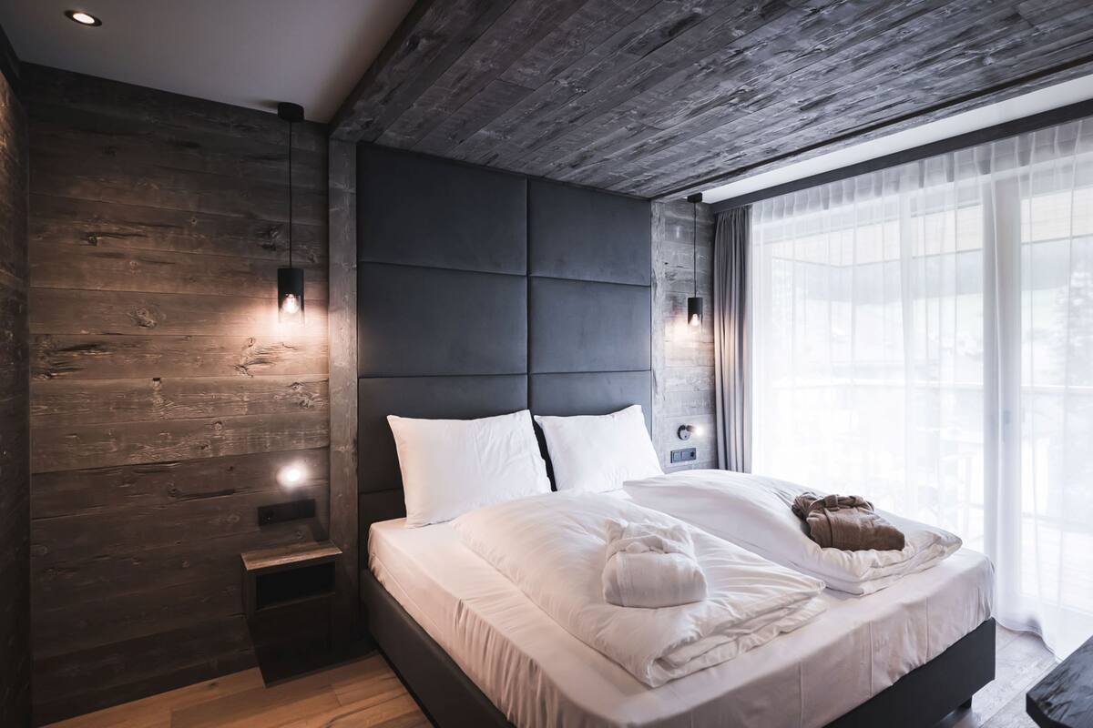 A modern bedroom with a large bed, wooden panel walls, and a large window with sheer curtains.