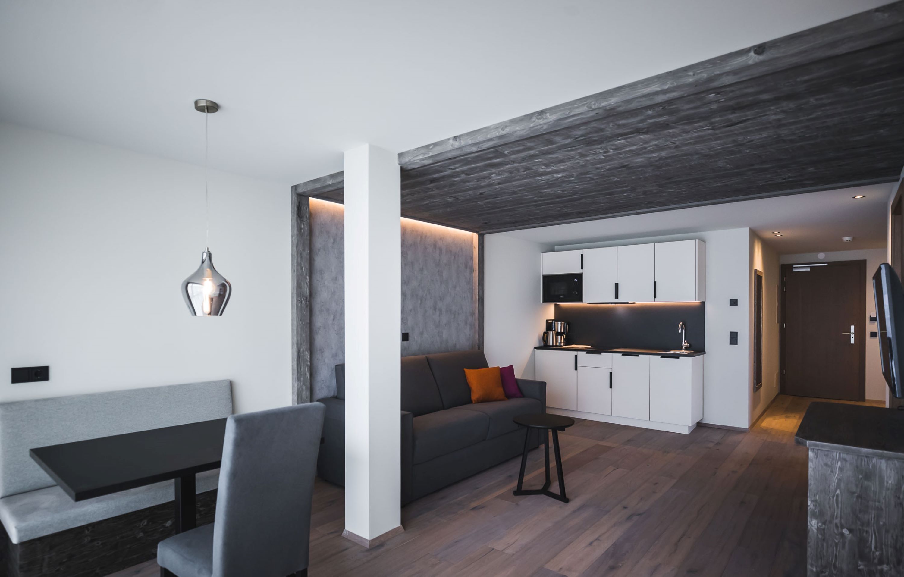 A modern living room with a grey sofa, a black table, and a small black stool. There is a kitchen area with white cabinets and a black countertop.