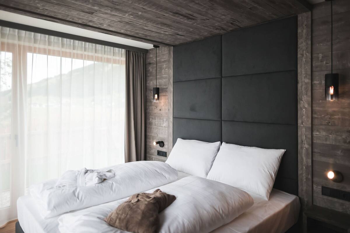A modern bedroom with a large bed, a headboard with a built-in light, and a view of the outdoors through sheer curtains.