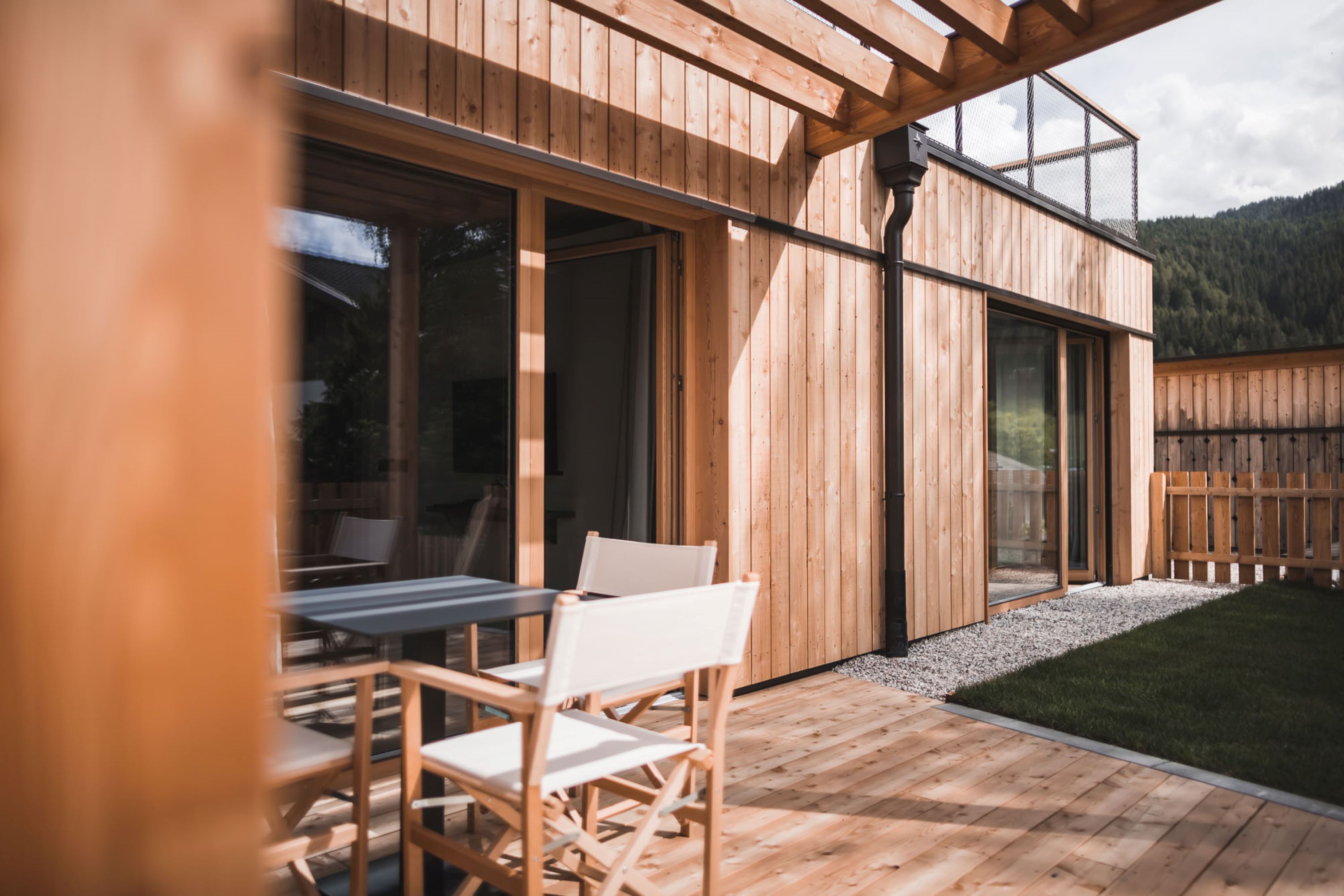 A wooden deck with a table and chairs, a wooden fence, and a view of a forested mountain.