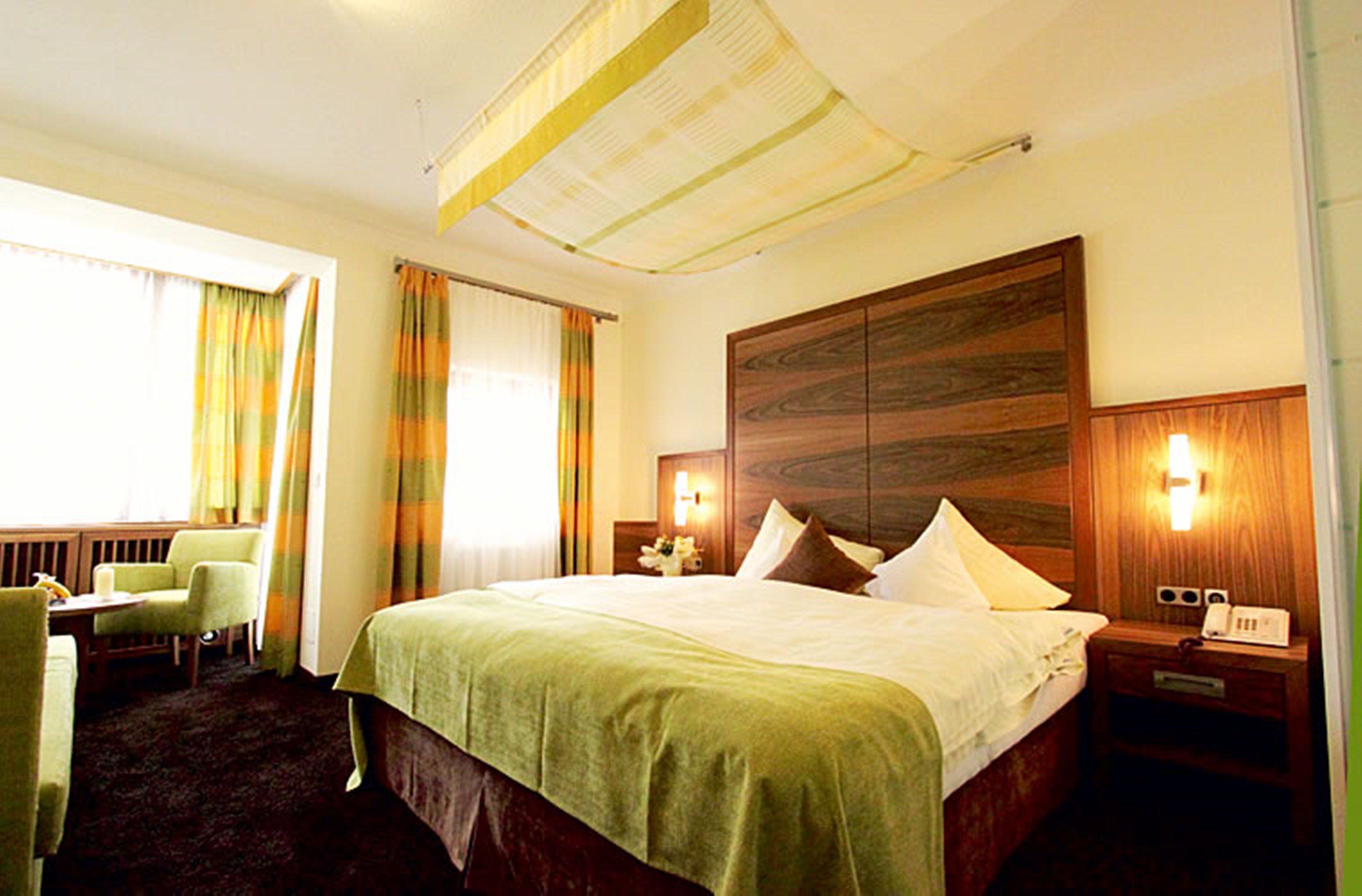 Quarto de hotel com uma cama grande, mesa de cabeceira, uma cadeira e cortinas.