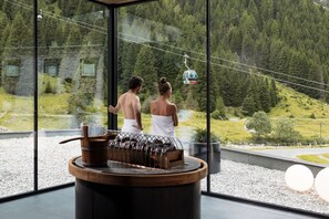 A modern spa area with a sauna, a glass wall offering a view of a forested mountain area, and a cable car in the background. - DAS VIER Sport,- und Wellnesshotel (Sankt Leonhard im Pitztal)