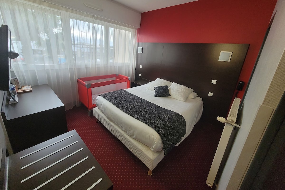 A hotel room with a bed, a desk, a chair, a red side table, and a window with sheer curtains.
