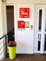 Entrada de un hotel con un letrero rojo que dice 'CIT' HOTEL, una placa con una 'H' grande y estrellas, una planta en maceta y una puerta con teclado y lector de tarjetas.