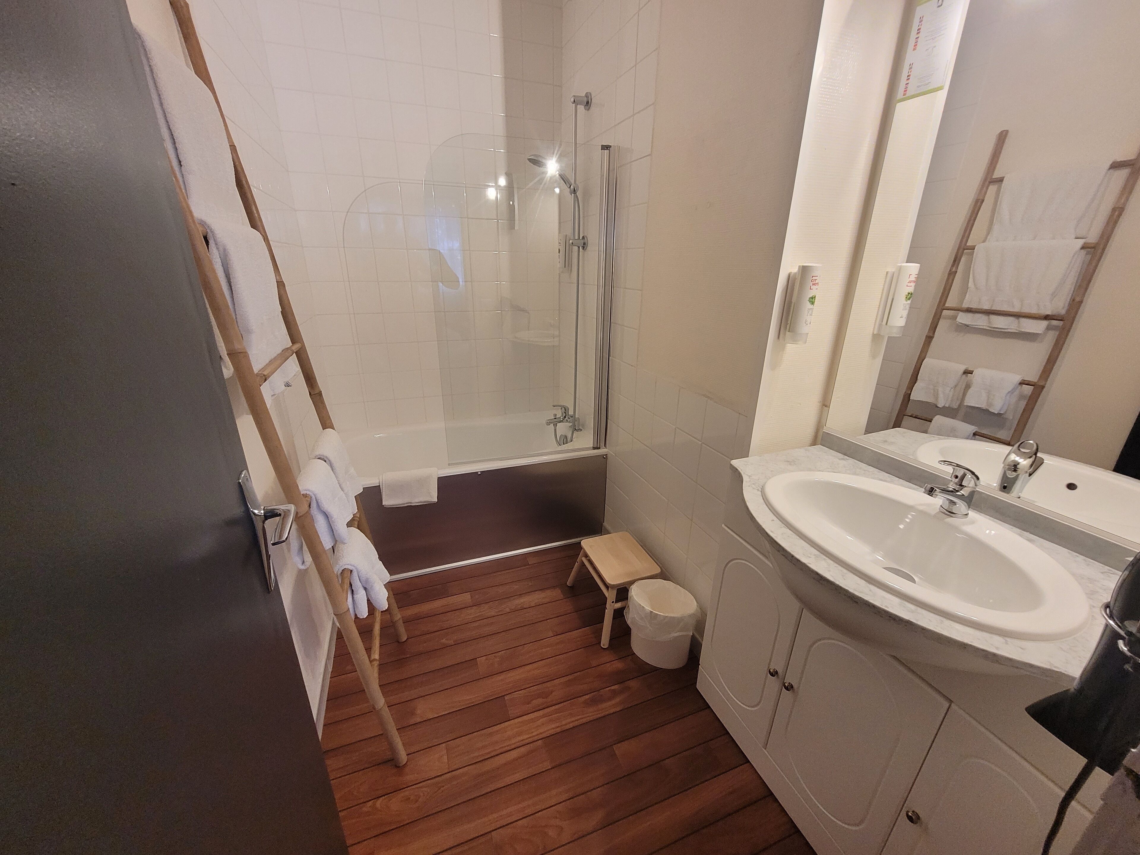 Ein modernes Badezimmer mit einer Glaskabine zur Dusche, Holzboden, weißem Waschbecken und einem Spiegel.