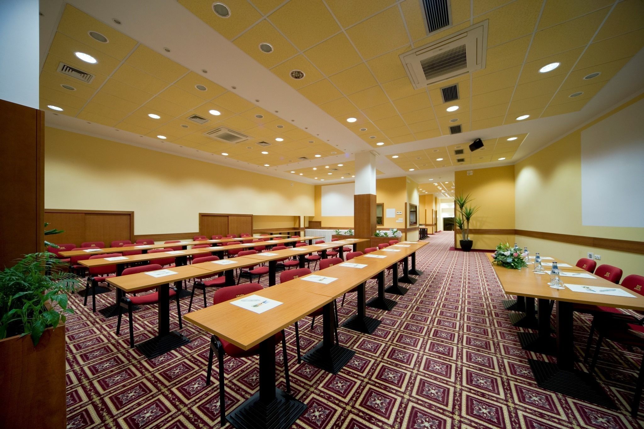 A conference room with rows of tables and chairs, a patterned carpet, and a yellow ceiling with recessed lighting.