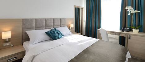 A hotel room with a bed, bedside table, desk, chair, and a potted orchid.