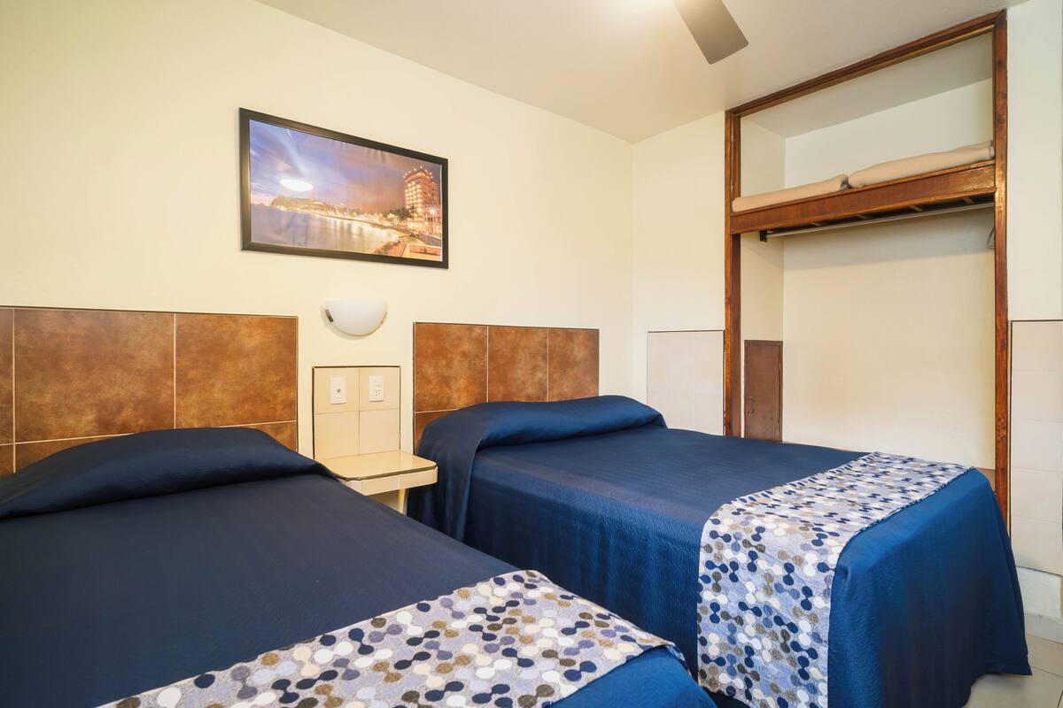 A hotel room with two beds, a nightstand, a wall-mounted lamp, and a cityscape painting.