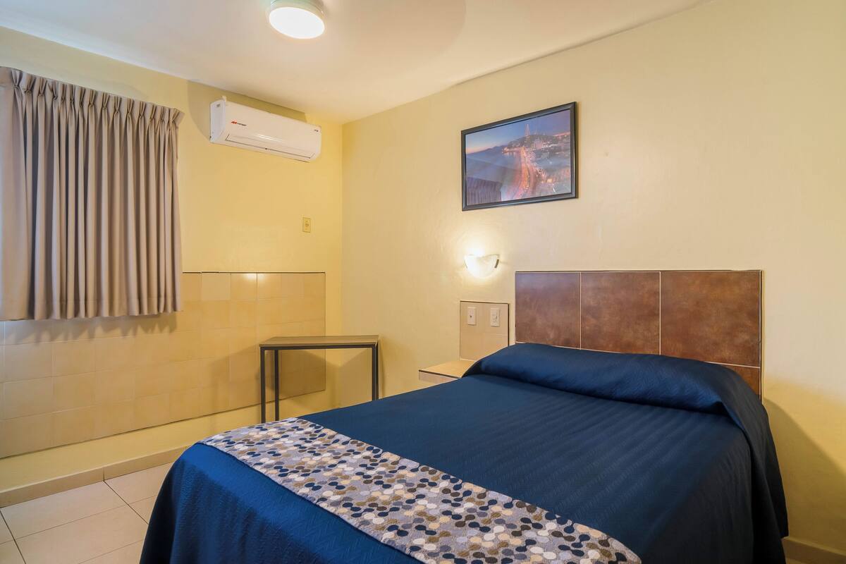 A hotel room with a bed, an air conditioning unit, a nightstand, a lamp, a picture on the wall, and a window with curtains.