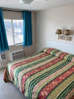 Un dormitorio con cama, ventana con cortinas, equipo de aire acondicionado y dos lámparas fijadas a la pared.