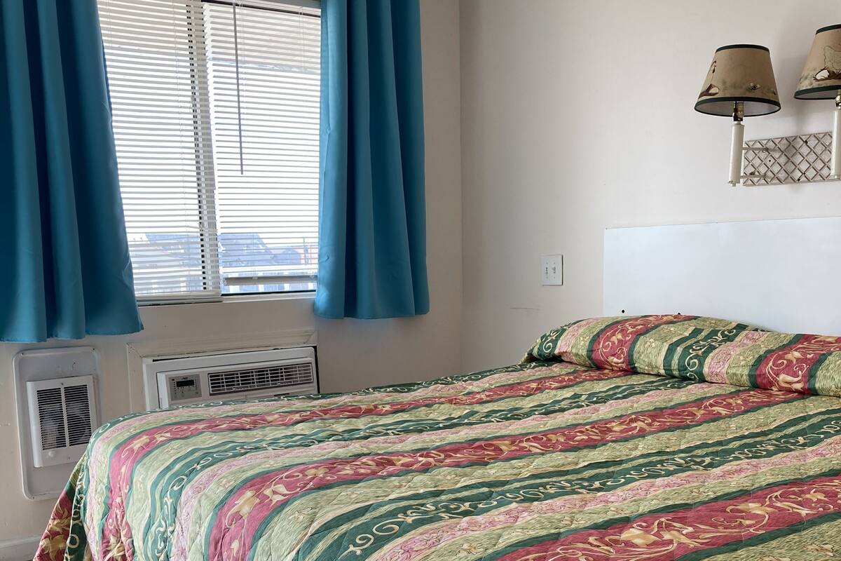 A bedroom with a bed, blue curtains, a window with blinds, a wall-mounted lamp, and a radiator.