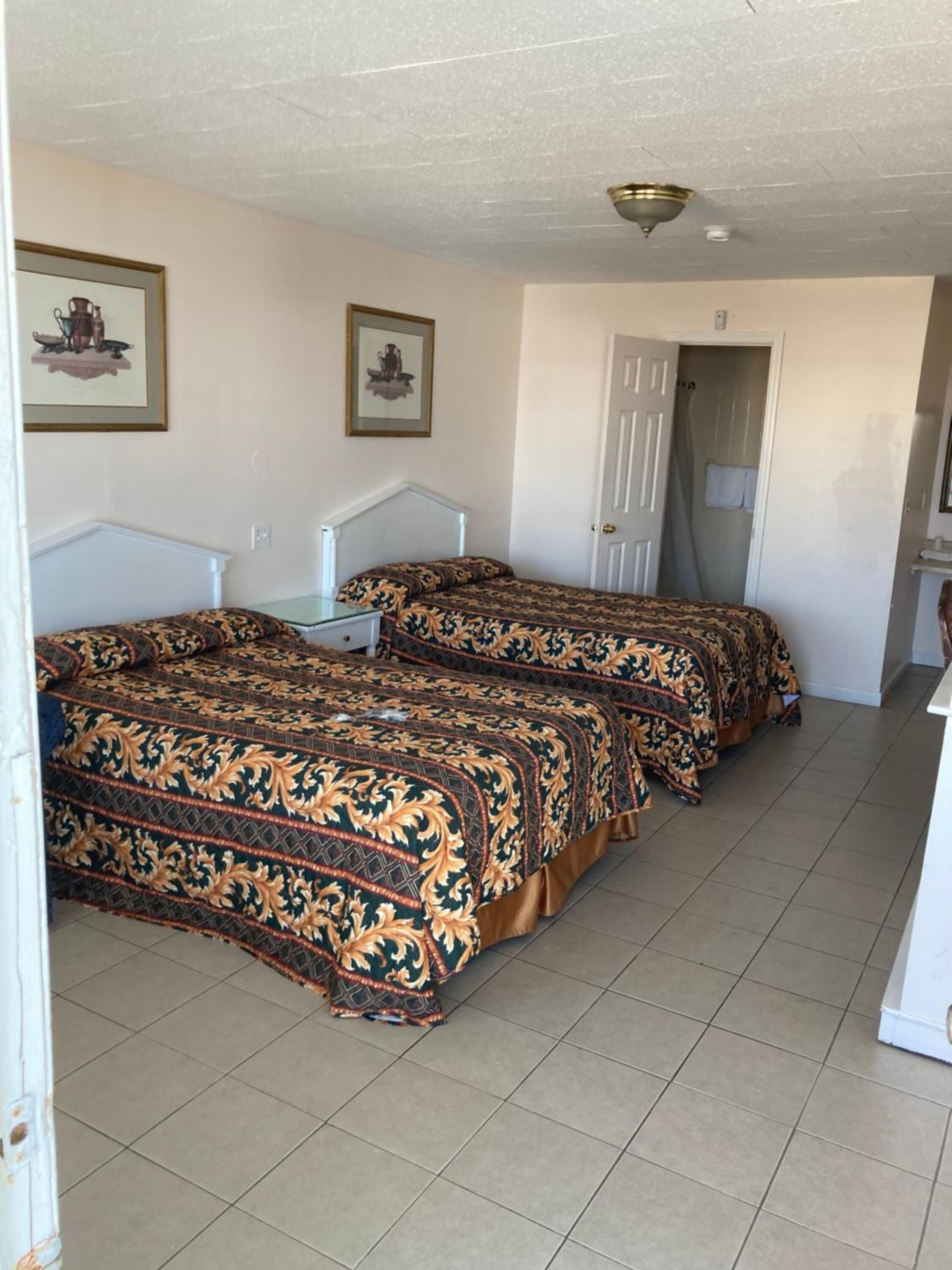 A room with two beds, each with a patterned bedspread, a white headboard, and a small table. There are framed pictures on the wall and a door leading to another room.