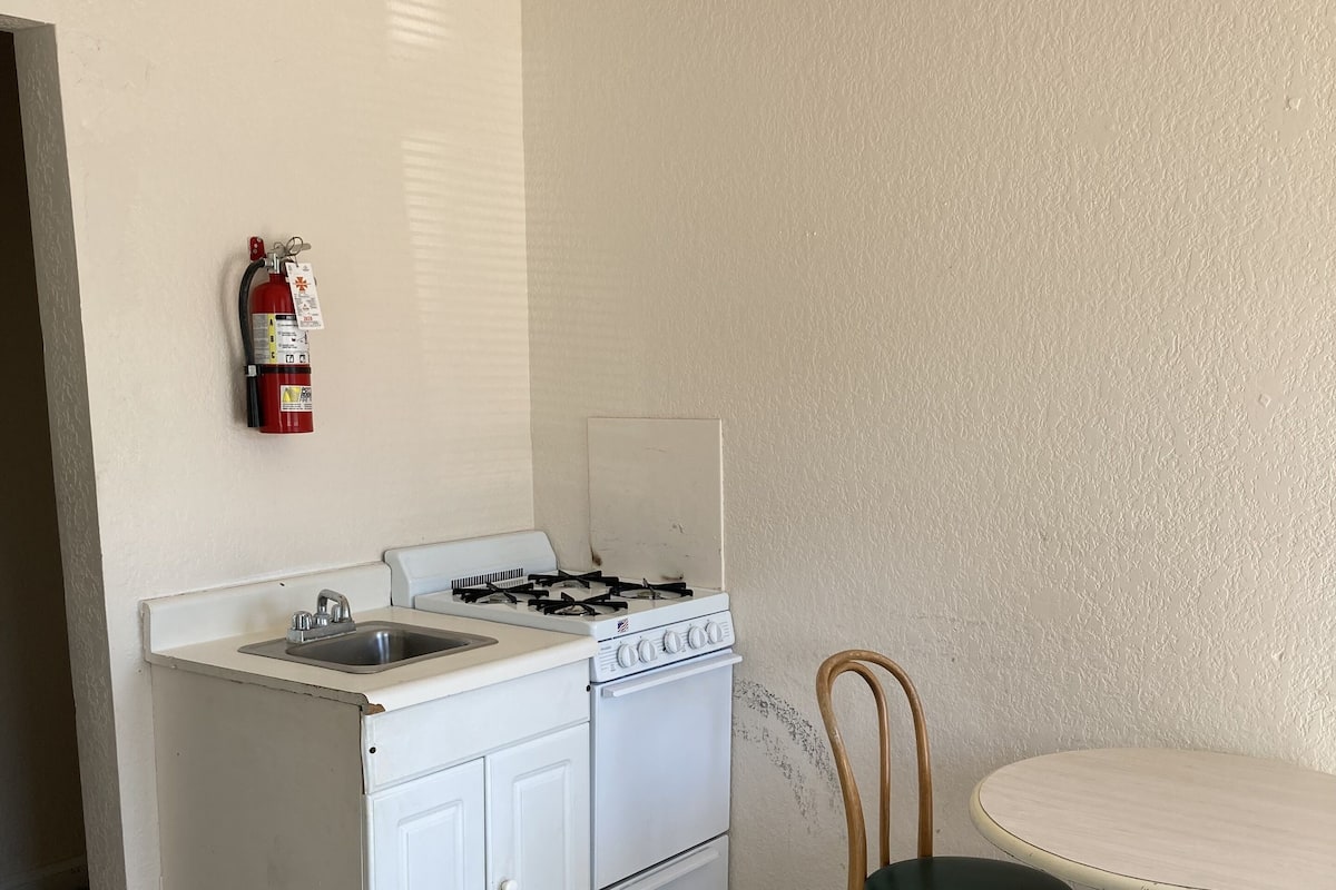 A compact kitchen with a white stove, sink, and cabinets, a wooden chair, and a small round table.