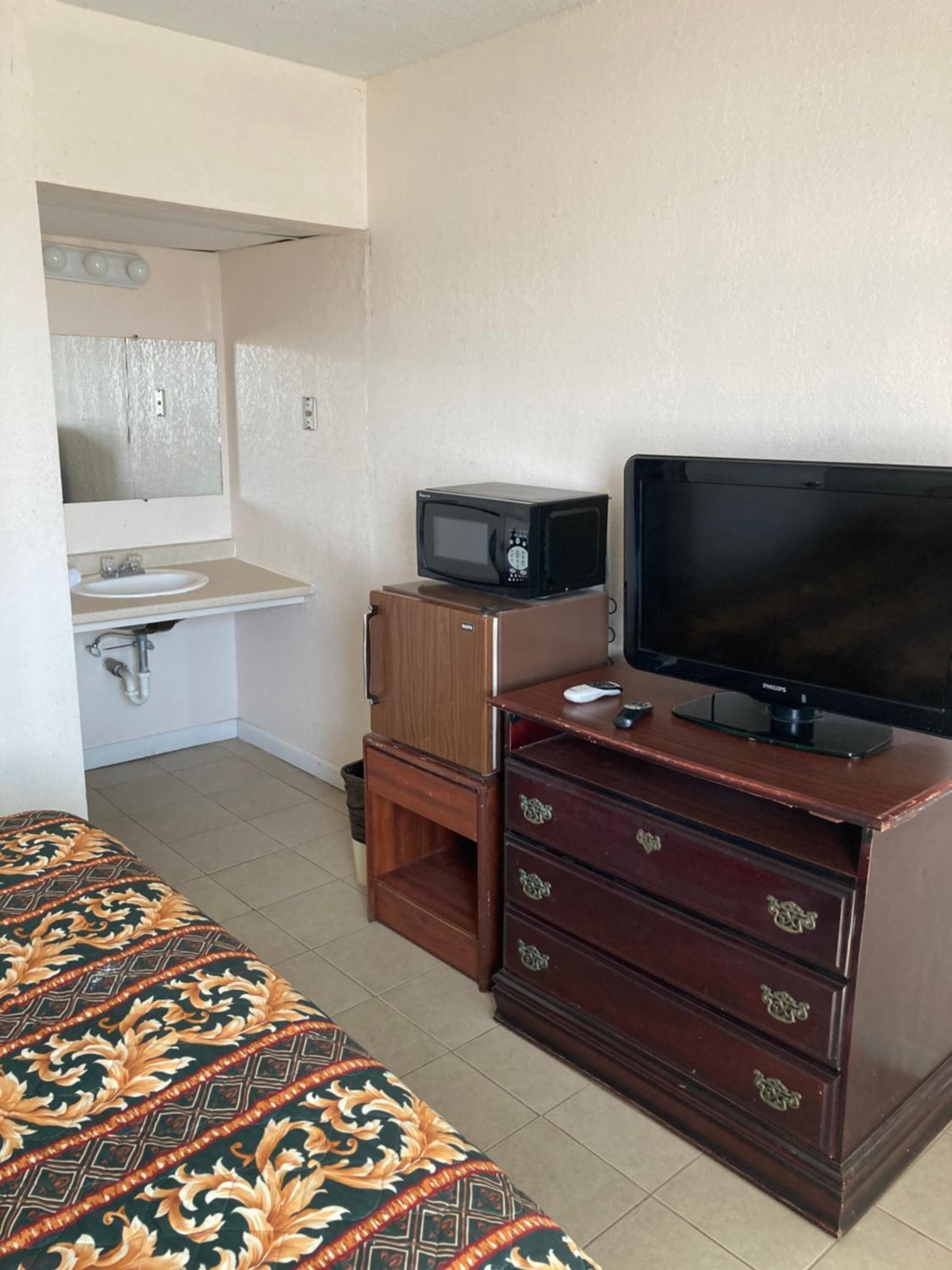 Una habitación de hotel compacta con una cama, un tocador de madera, un televisor, un microondas y un lavabo.