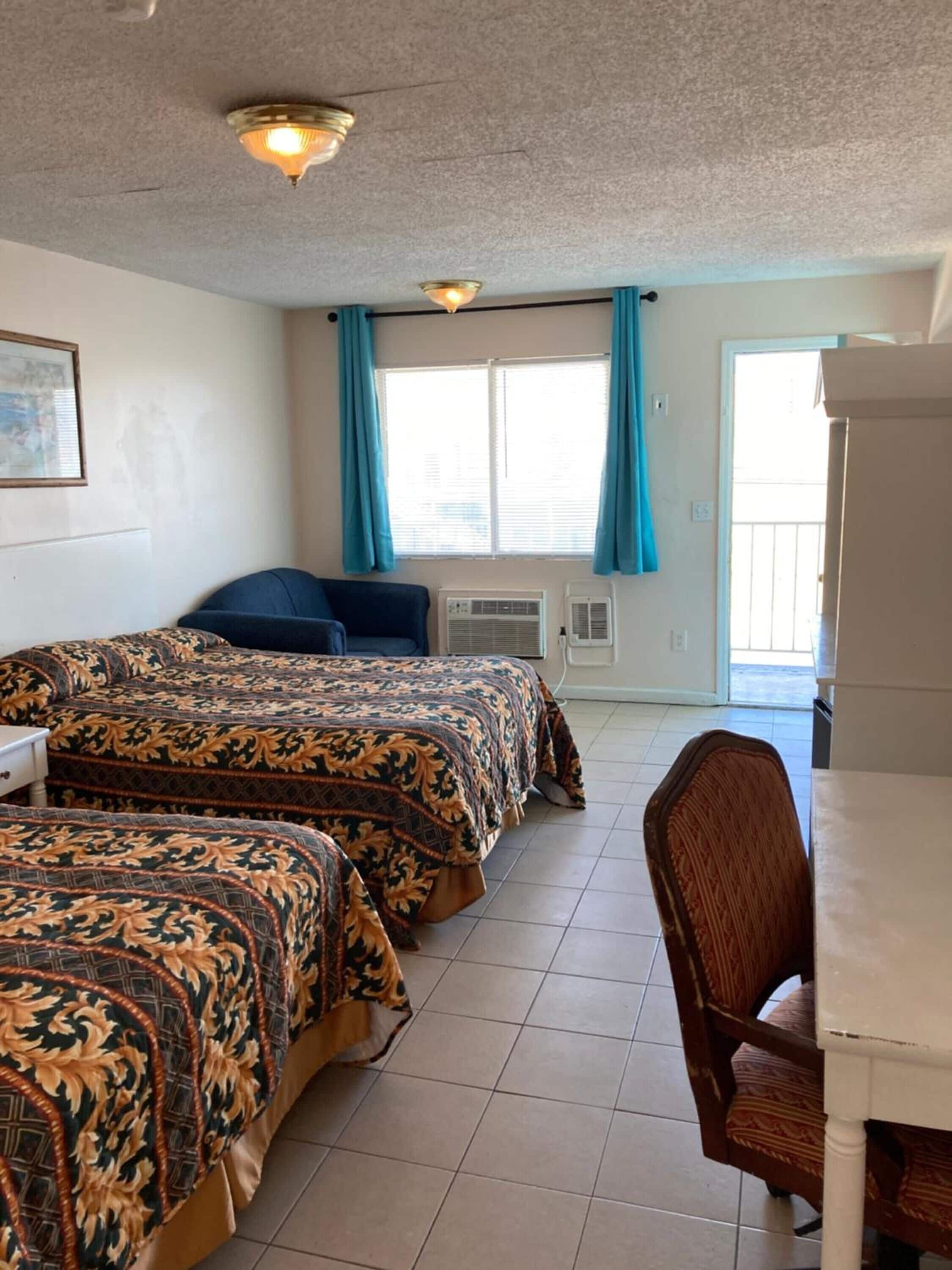 A room with two beds, a desk, and a chair. There is a window with curtains, an air conditioning unit, and a ceiling light fixture.