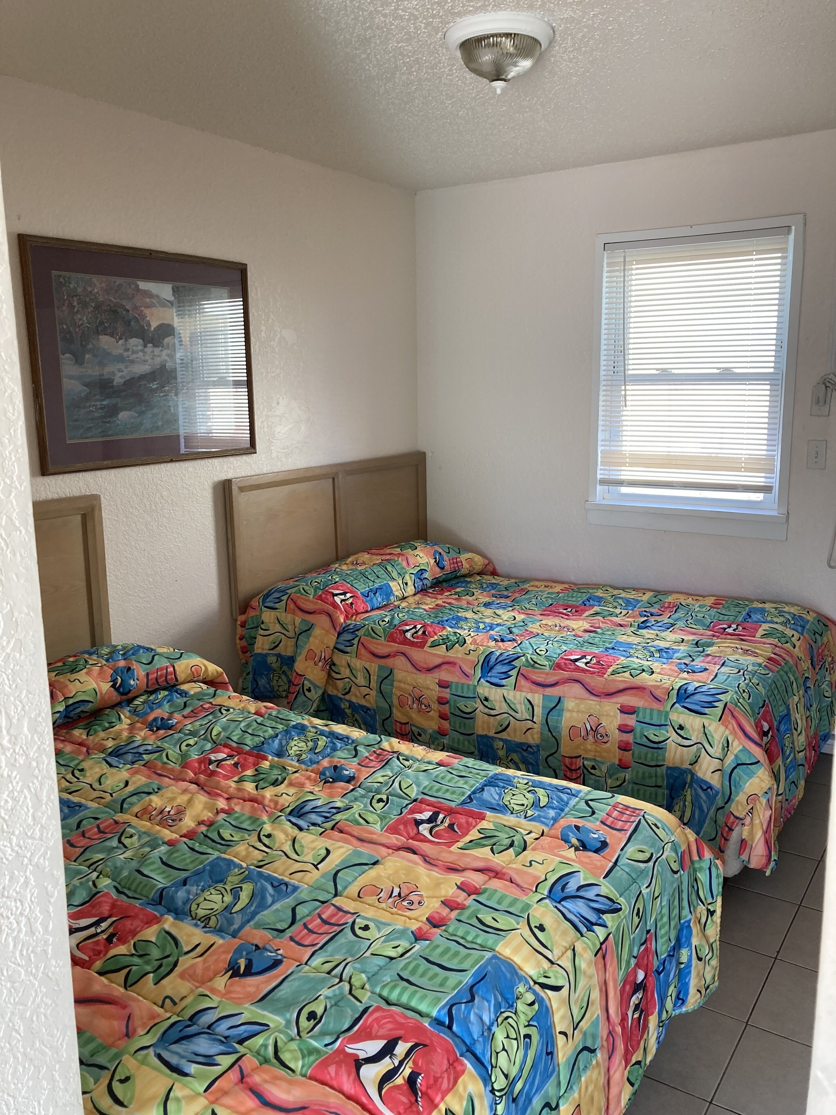 A room with two beds, a ceiling light, a window with blinds, and a framed picture on the wall.