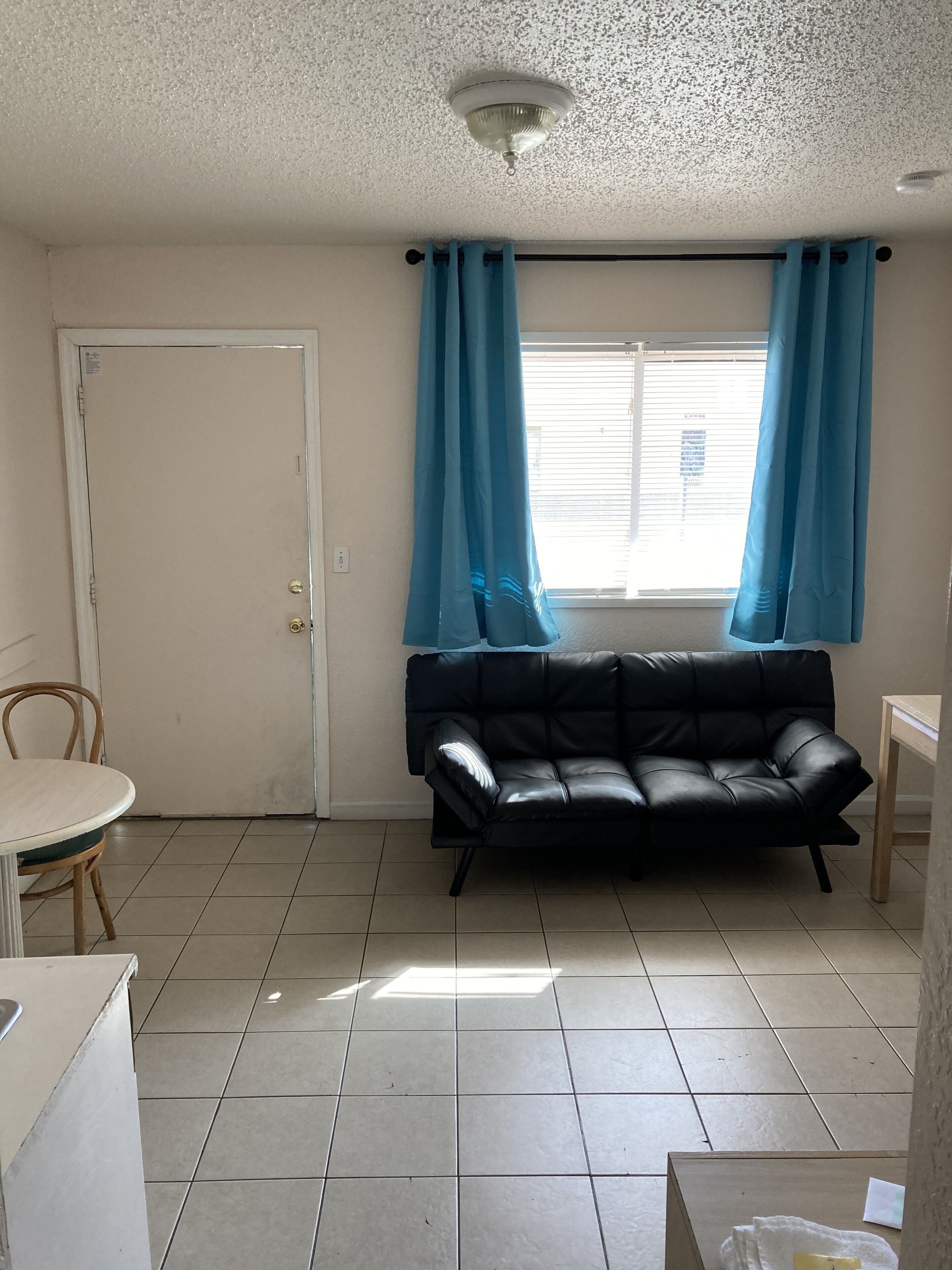 A small, sparsely furnished room with a black leather sofa, a wooden table, and a chair. A door and a window with blue curtains are visible.