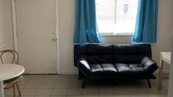 A small, sparsely furnished room with a black leather sofa, a wooden table, and a chair. A door and a window with blue curtains are visible.