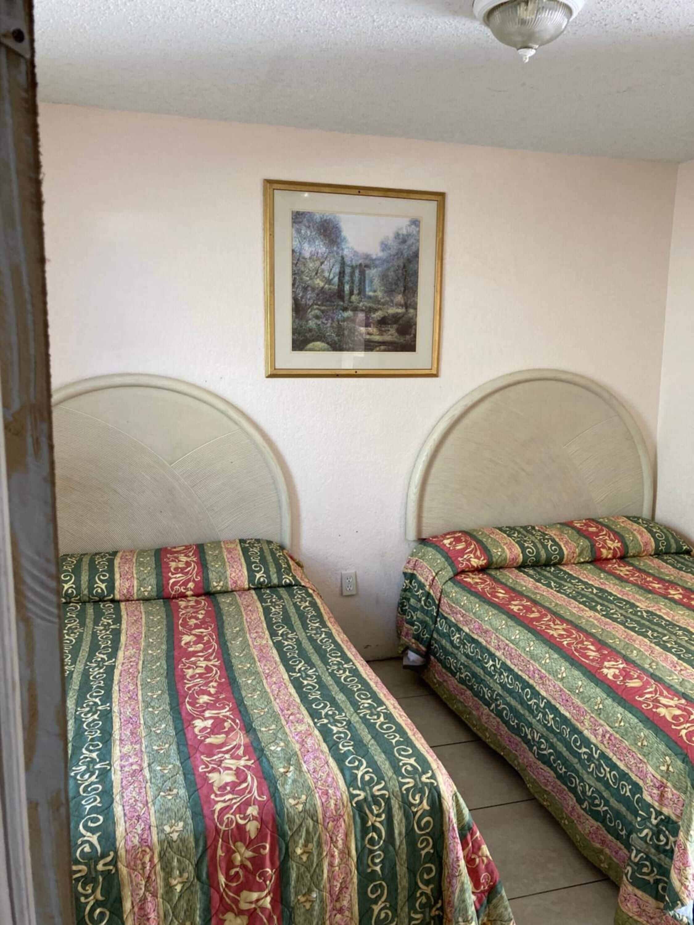 Ein Zimmer mit zwei Betten, jeweils mit gemusterten Bettdecken. Darüber an der Wand hängt ein gerahmtes Bild.