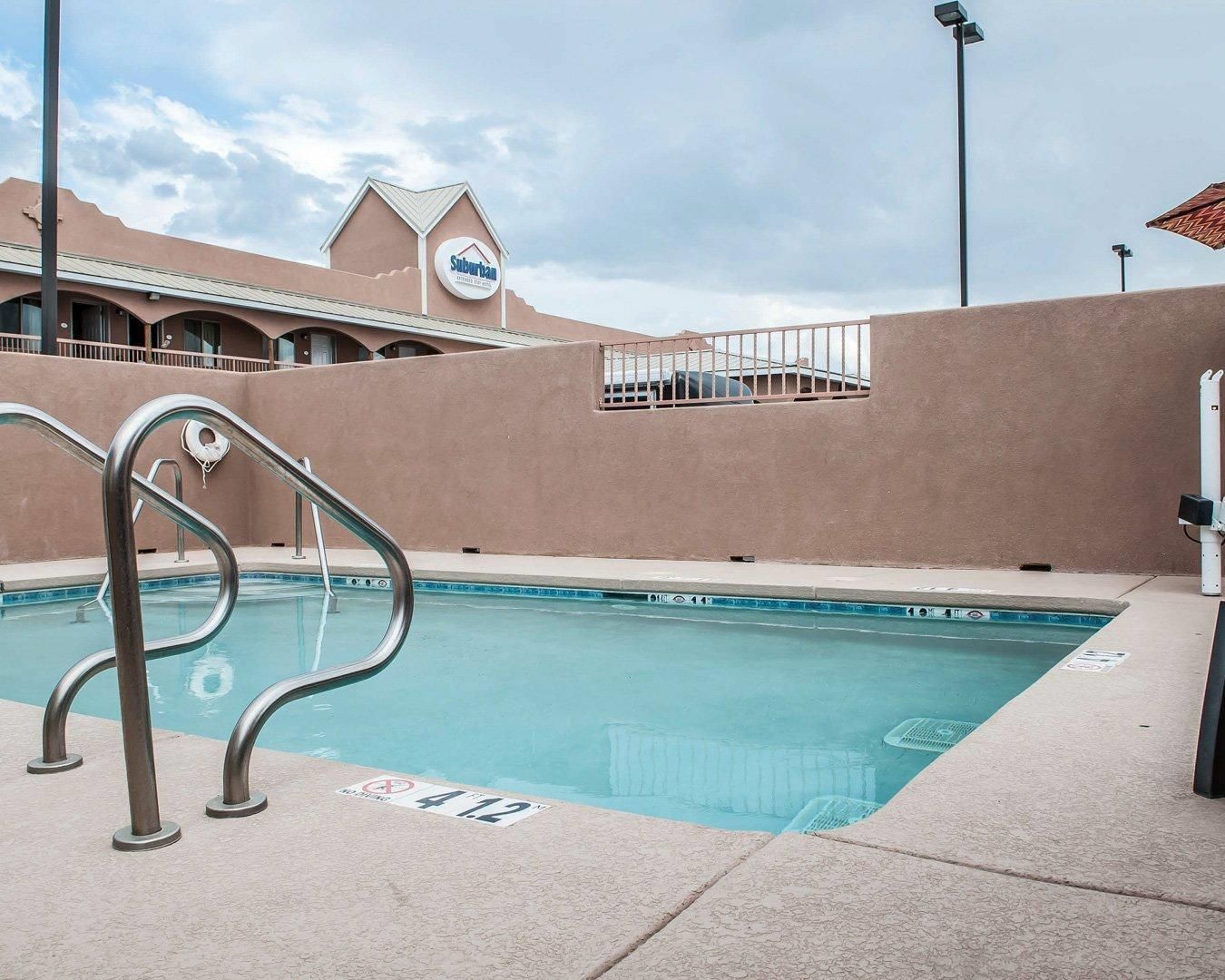 Una piscina con barandillas de metal, un cartel de Suburban y un edificio con un estilo arquitectónico particular al fondo.