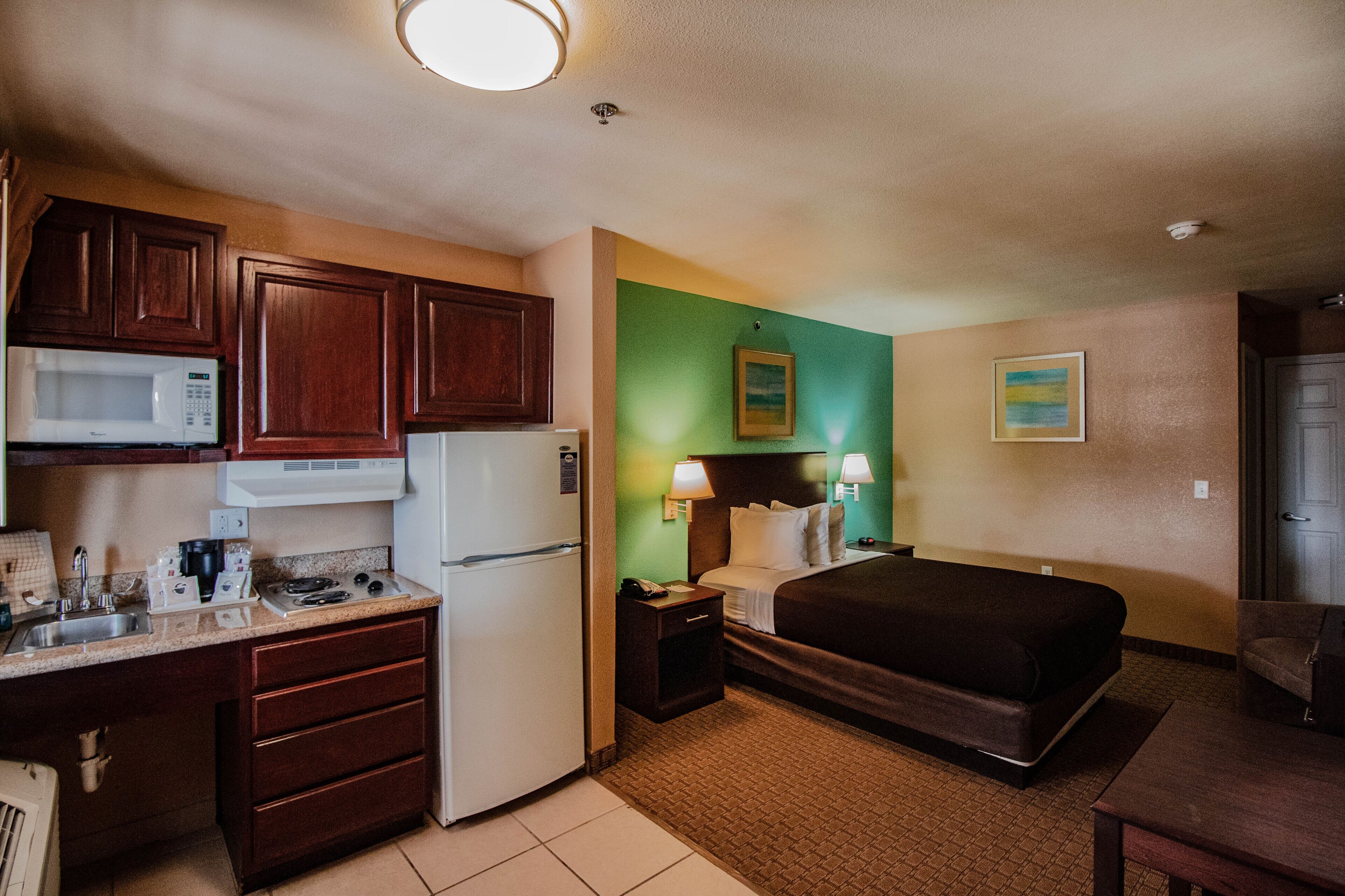 A compact hotel room with a kitchenette, a bed, a bedside table, and a nightstand.
