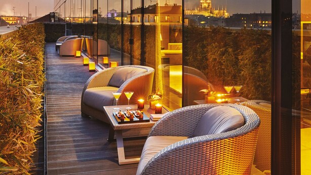 Un espacio moderno al aire libre con sillas de mimbre y una mesa, con vista a un paisaje urbano al anochecer.
