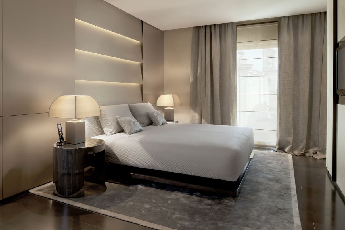 A modern bedroom with a large bed, bedside lamps, and a view of the city through curtains.
