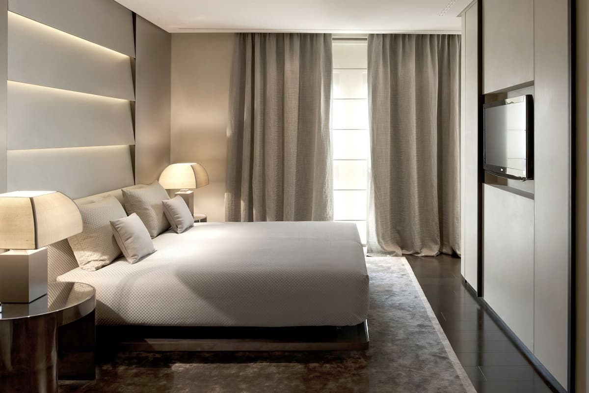 A modern hotel room with a large bed, bedside lamps, a flat-screen TV, and a large window with curtains.