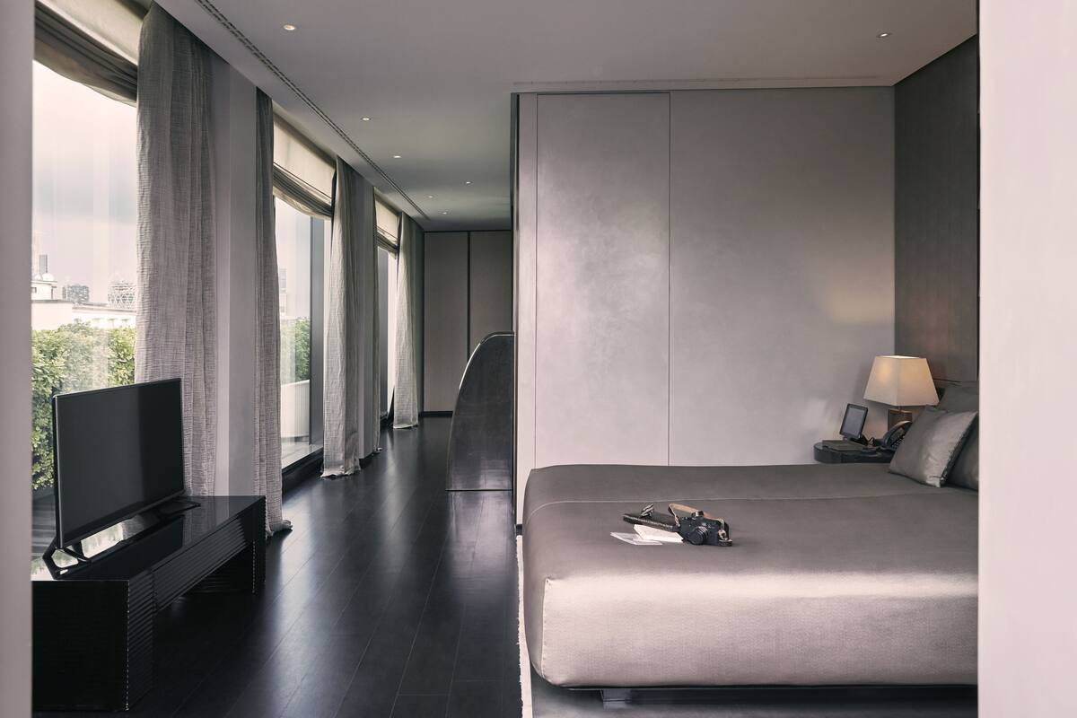 A modern hotel room with a large bed, a flat-screen TV on a black stand, a desk with a lamp, and a view of the city through large windows.