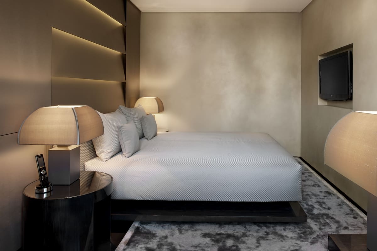 A modern hotel room with a large bed, two bedside tables with lamps, a flat-screen TV mounted on the wall, and a marble-patterned carpet.