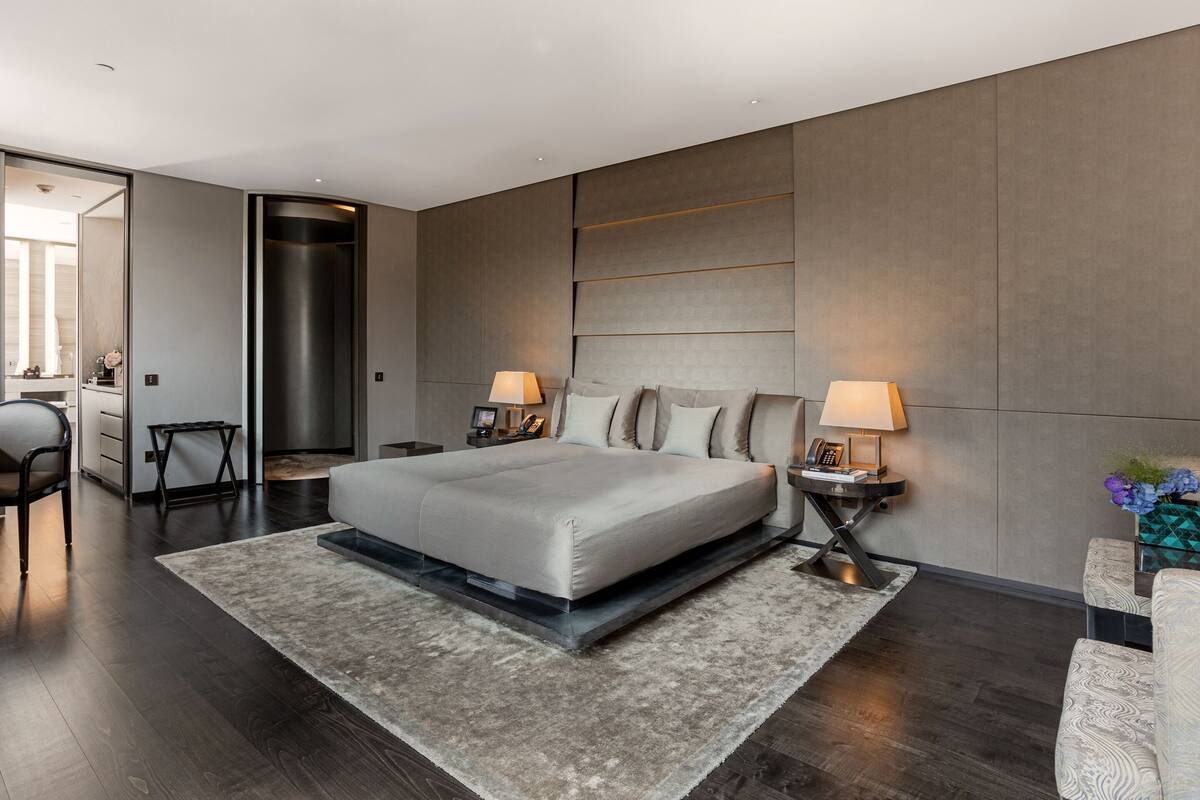 A modern bedroom with a large bed, two bedside tables with lamps, a built-in wardrobe, and a dark wood floor.